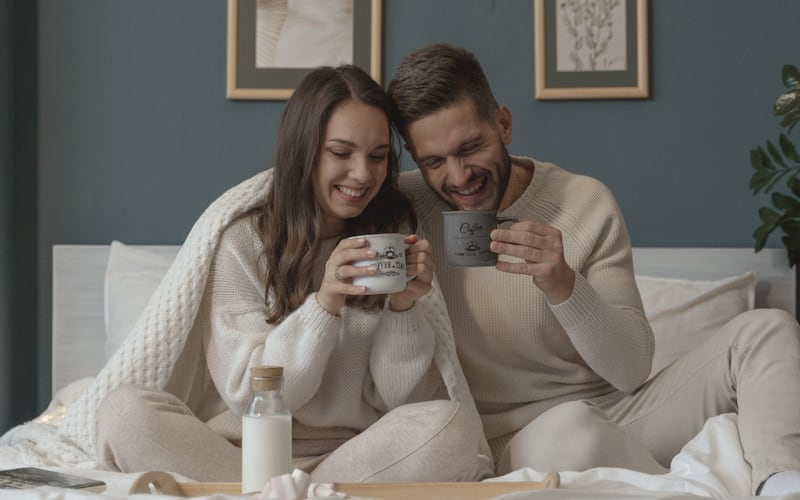 Una pareja vestida de blanco sobre una cama tomando desayuno. Ambos tienen un tazón en la mano.