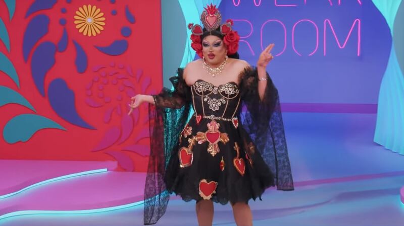 Drag con maquillaje recargado, con vestido negro hasta la mitad del muslo y flores rojas en varias partes, lleva una corona de colores, en un traje flocklórico mexicano