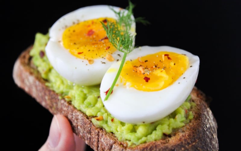 Sobre fondo negro, primer plano de una mano sosteniendo una tostada de pan integral con palta y huevos.