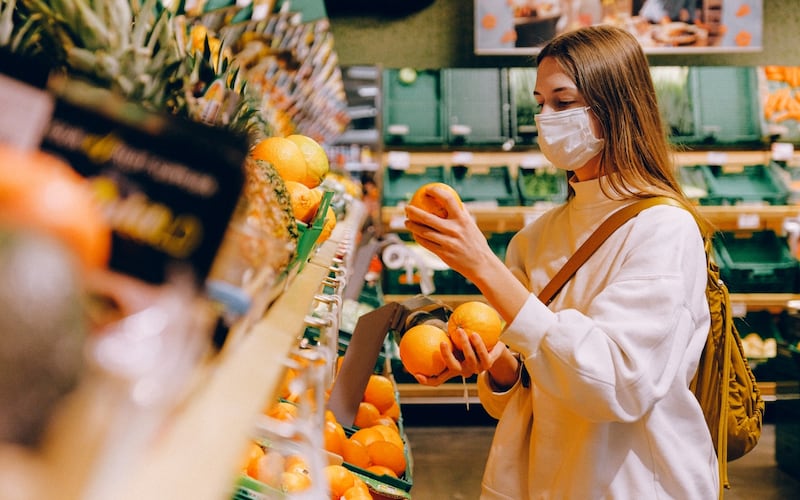 Una mujer joven vestida de blanco compra en el supermercado. Sostiene tres naranjas en sus manos.