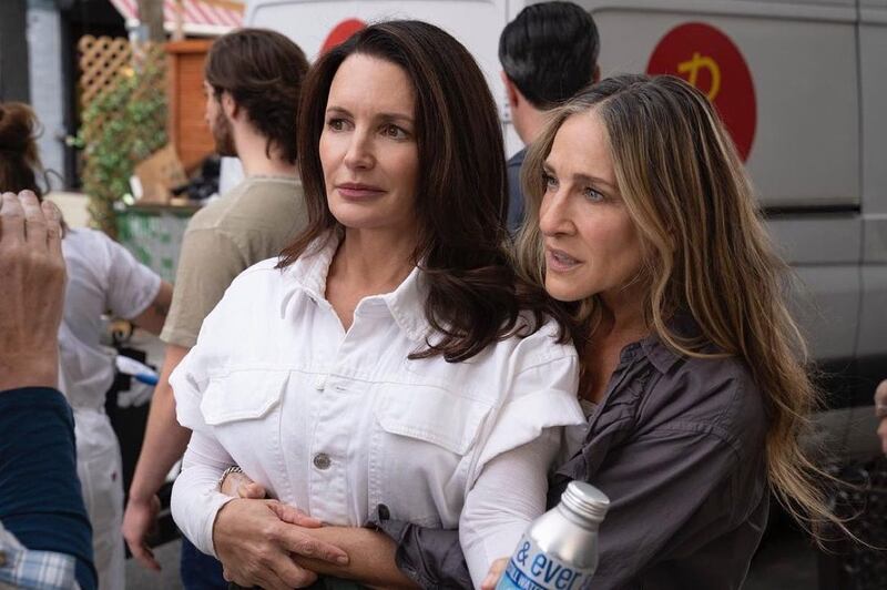 Kristin Davis, con una blusa blanca, es tomada por la cintura por Sarah Jessica, que va con una blusa gris oscuro, como deteniéndola, mientras están en una calle y se ven vehículos atrás.